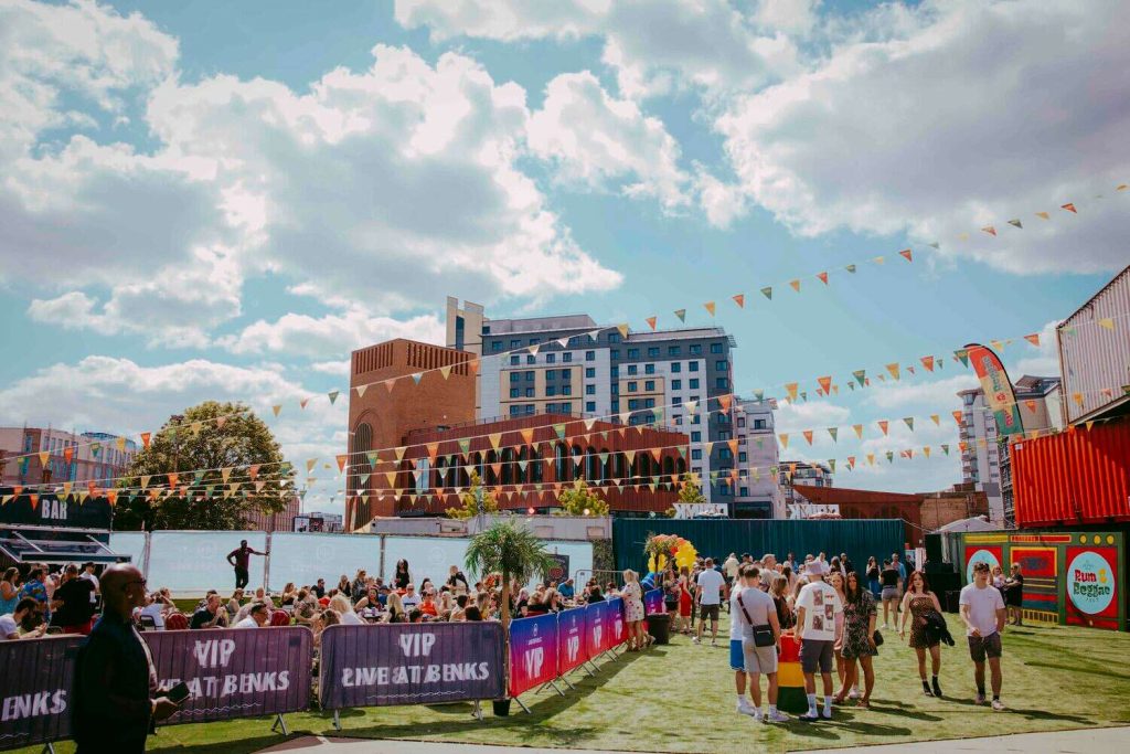 Blackscarletclubs_A-3473 A lively outdoor event with people mingling under a blue sky with clouds. Theres a bar area on the left, vibrant flags strung above, and colorful shipping containers around. Buildings are visible in the background.