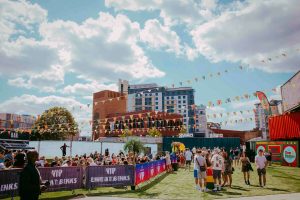 A lively outdoor event with people mingling under a blue sky with clouds. Theres a bar area on the left, vibrant flags strung above, and colorful shipping containers around. Buildings are visible in the background.