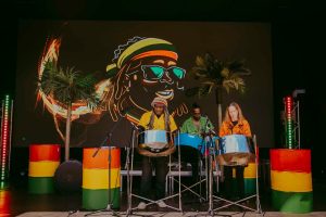 Three musicians playing steel drums stand on stage. The backdrop features a vibrant illustration of a person with sunglasses and a hat. Palm trees and colorful lights accentuate the lively atmosphere.