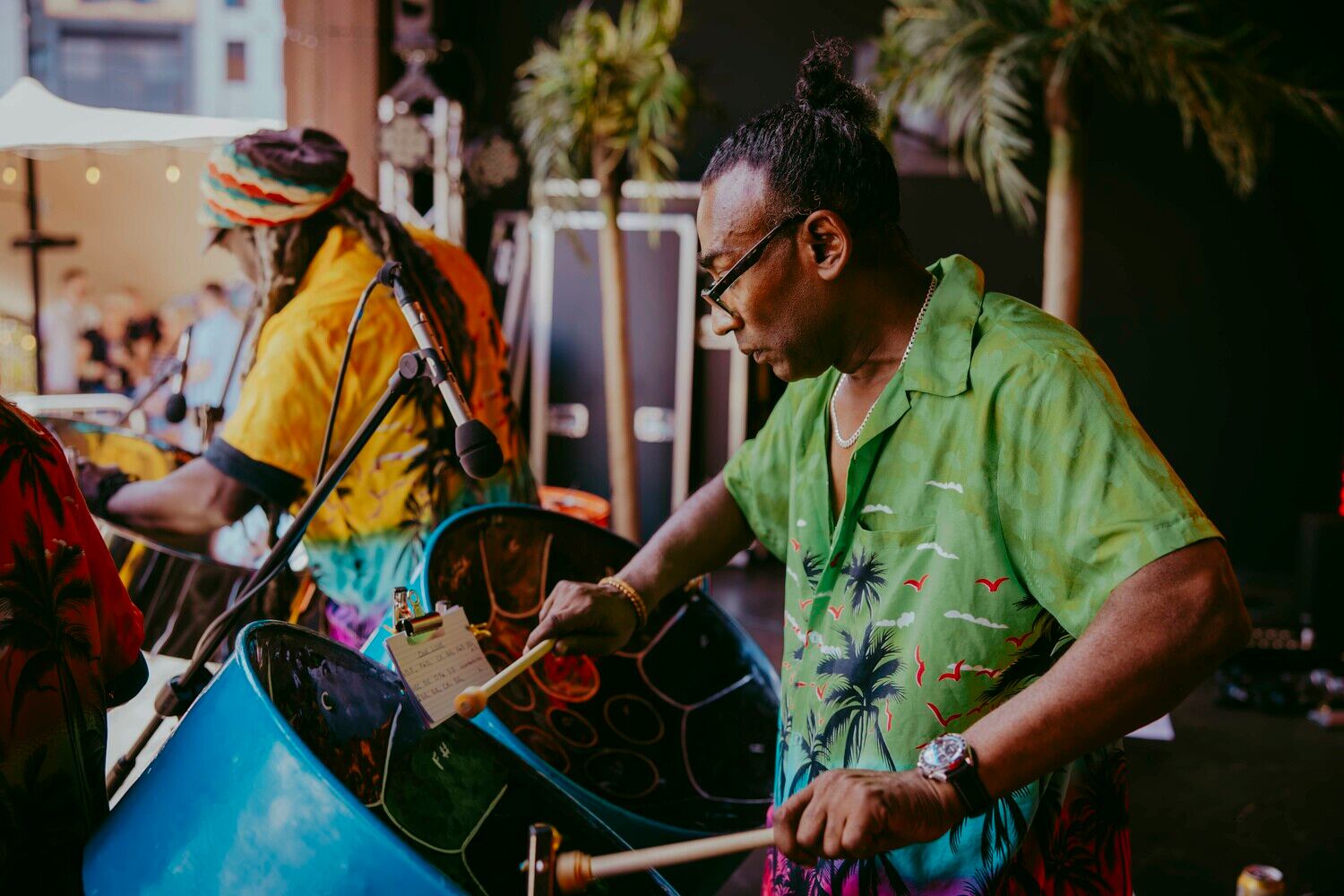 Two musicians play steel drums on stage. The nearest musician wears a green tropical shirt and glasses, while the other wears a yellow shirt and multicolored cap. Palm plants are visible in the background.
