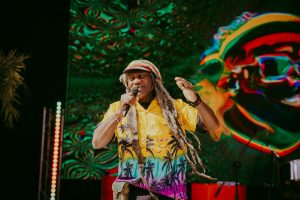 Man with long hair in a colorful shirt and hat performs on stage with a microphone. A vibrant, psychedelic design is displayed on a large screen behind him. Musical equipment is visible in the background.