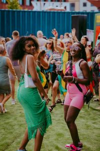 Two women smiling and posing at an outdoor event. One wears a green dress and the other a pink outfit. People in the background are enjoying the event, holding drinks, with a blue shipping container visible. The atmosphere is lively and festive.