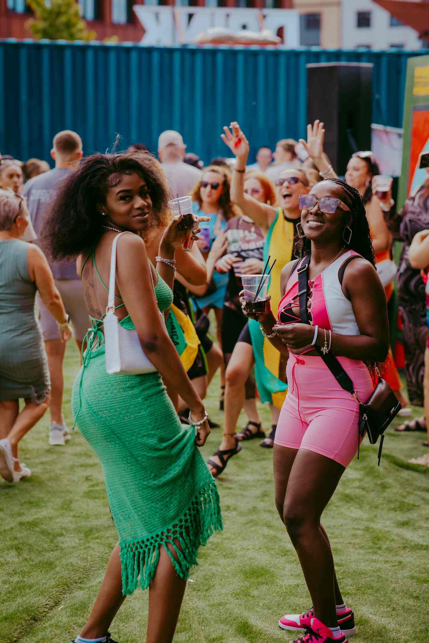 Two women smiling and posing at an outdoor event. One wears a green dress and the other a pink outfit. People in the background are enjoying the event, holding drinks, with a blue shipping container visible. The atmosphere is lively and festive.