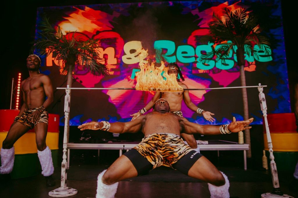 Blackscarletclubs_A-3684 Three performers dressed in animal print shorts and leg warmers are participating in a limbo contest. The central figure is leaning back under a bar with a flame on it. Vibrant colors and palm trees are in the background.
