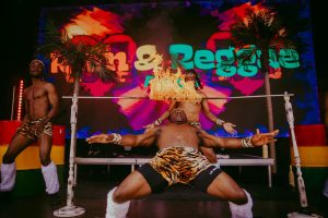 Three performers dressed in animal print shorts and leg warmers are participating in a limbo contest. The central figure is leaning back under a bar with a flame on it. Vibrant colors and palm trees are in the background.