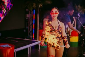 A performer wearing a flame-patterned outfit holds a lit fire staff, with vibrant stage lights and palm decorations in the background. Another person is visible playing guitar. The scene conveys a lively, tropical atmosphere.