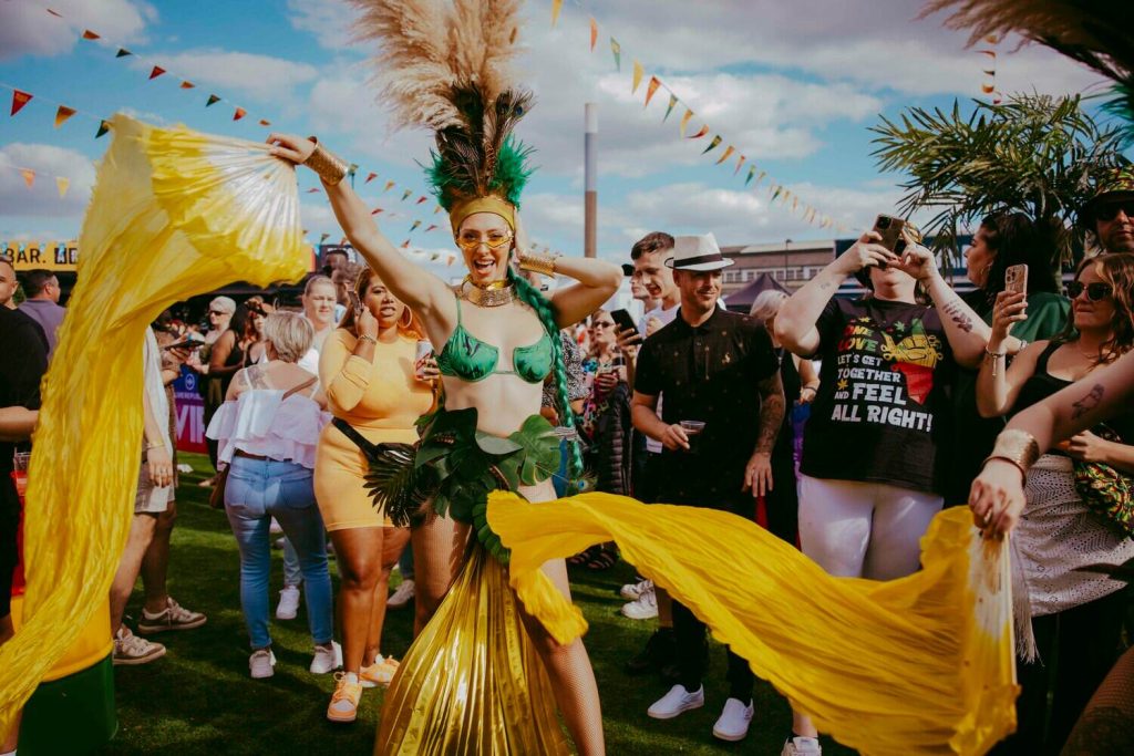 Blackscarletclubs_A-3838 A lively outdoor festival scene with a woman in a vibrant costume dancing with yellow fabric. She wears a green outfit and a feathered headdress. Crowds of people watch and enjoy the event under a partly cloudy sky with festive bunting overhead.