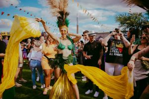 A lively outdoor festival scene with a woman in a vibrant costume dancing with yellow fabric. She wears a green outfit and a feathered headdress. Crowds of people watch and enjoy the event under a partly cloudy sky with festive bunting overhead.