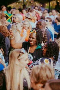 A woman smiling and holding a small Jamaican flag stands in a lively crowd. She wears a colorful headband and black top. The atmosphere is festive, with people of various backgrounds enjoying the event under warm lighting.