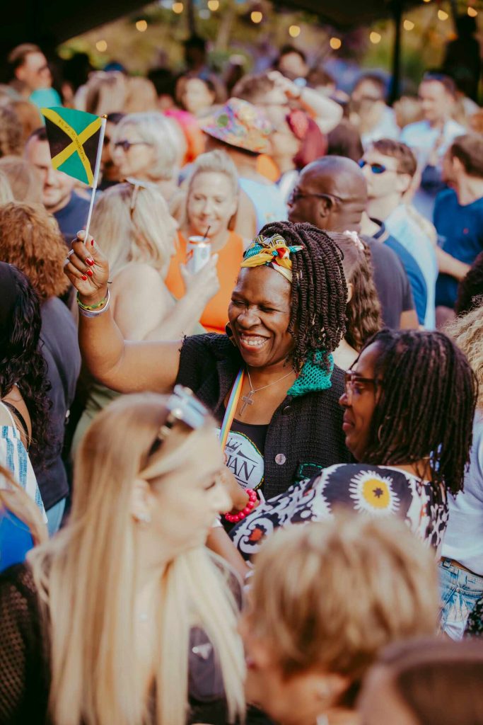 Blackscarletclubs_A-3913 A woman smiling and holding a small Jamaican flag stands in a lively crowd. She wears a colorful headband and black top. The atmosphere is festive, with people of various backgrounds enjoying the event under warm lighting.