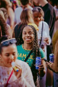 A young woman with long braided hair smiles while holding a drink can in a crowded outdoor gathering. She wears a green shirt with yellow lettering. People around her are also holding drinks, and one person blows a party horn.
