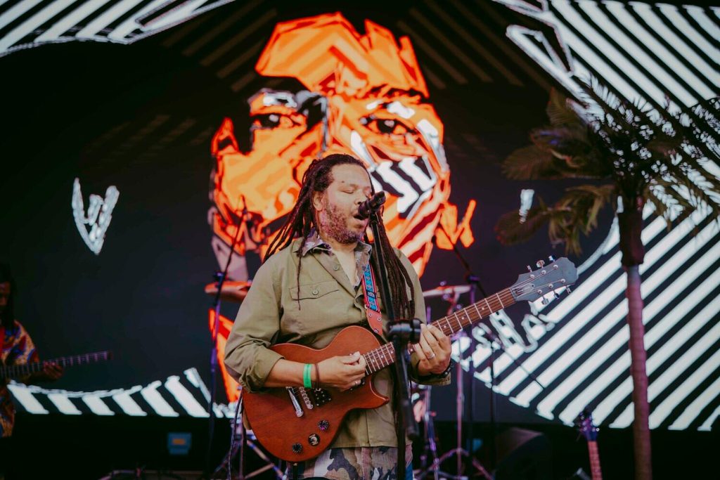 Blackscarletclubs_A-4056 A musician with long hair plays an electric guitar and sings into a microphone on stage. Behind him is a large LED screen displaying an abstract face design in orange and white. A small palm tree is visible to the side.