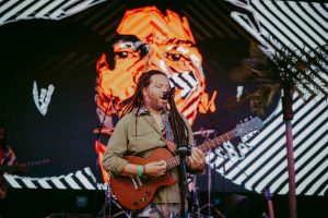 A musician with long hair plays an electric guitar and sings into a microphone on stage. Behind him is a large LED screen displaying an abstract face design in orange and white. A small palm tree is visible to the side.