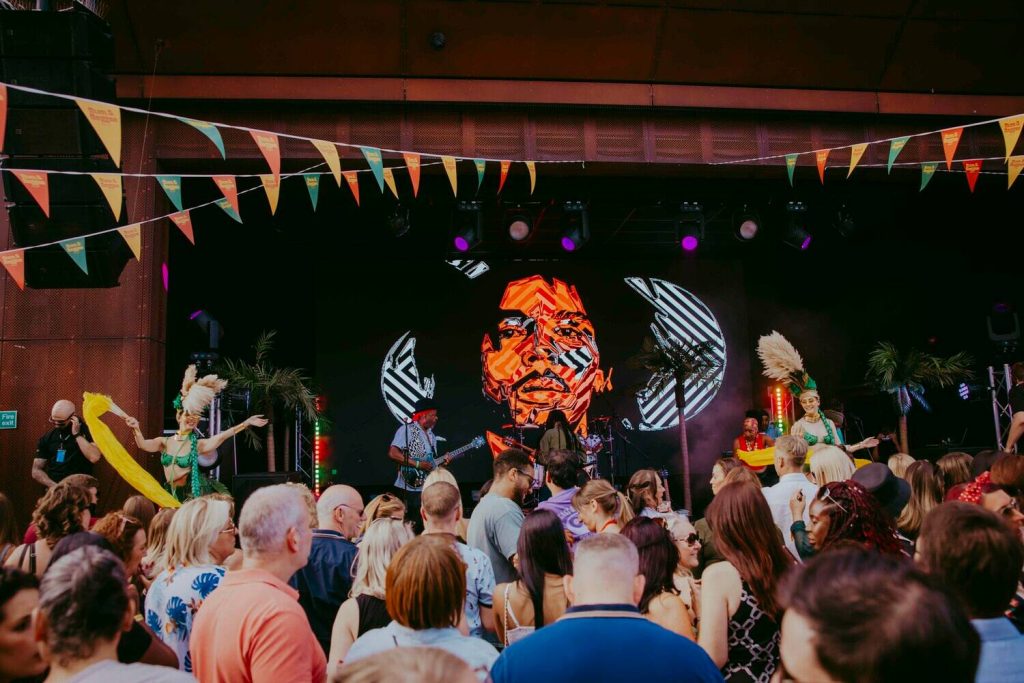 A lively crowd enjoys a vibrant outdoor concert. Performers in colorful costumes dance on stage with a vivid digital art backdrop. Multicolored flags hang above, enhancing the festive atmosphere.