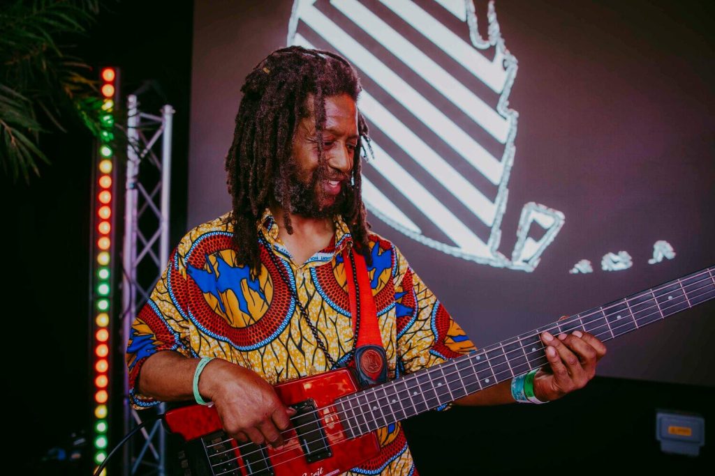 A musician with long dreadlocks plays a bass guitar on stage. He wears a colorful, patterned shirt and wristbands. Behind him is a large screen with abstract graphics and a lighting rig with multicolored lights.