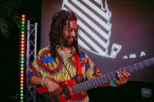 A musician with long dreadlocks plays a bass guitar on stage. He wears a colorful, patterned shirt and wristbands. Behind him is a large screen with abstract graphics and a lighting rig with multicolored lights.