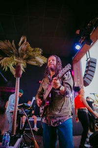 Musician with dreadlocks playing guitar on stage under palm tree, wearing a green shirt. Other band members and instruments visible, including a keyboard and microphone. Stage lighting creates a vibrant atmosphere.