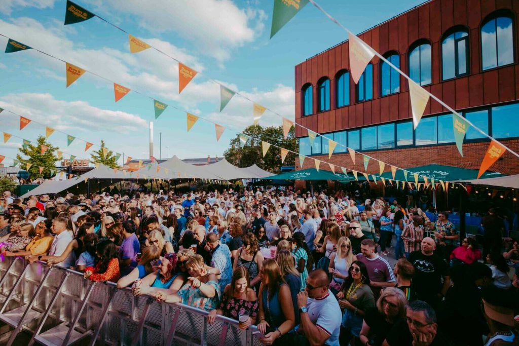 Blackscarletclubs_A-4142 A lively outdoor music festival with a large crowd gathered in front of a stage. Colorful triangular banners hang overhead. A modern brick building is in the background. The sky is partly cloudy, and people are enjoying the event.