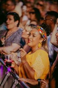 A woman in a yellow top and colorful headband holds a drink and looks toward a performance. She is in a crowd at an outdoor event, under warm lighting. Other people around her are watching the show and taking photos.