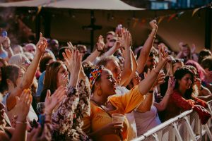 A diverse crowd of people enthusiastically cheer and raise their hands at an outdoor event, under a canopy with string lights and colorful bunting. The atmosphere is lively and festive.