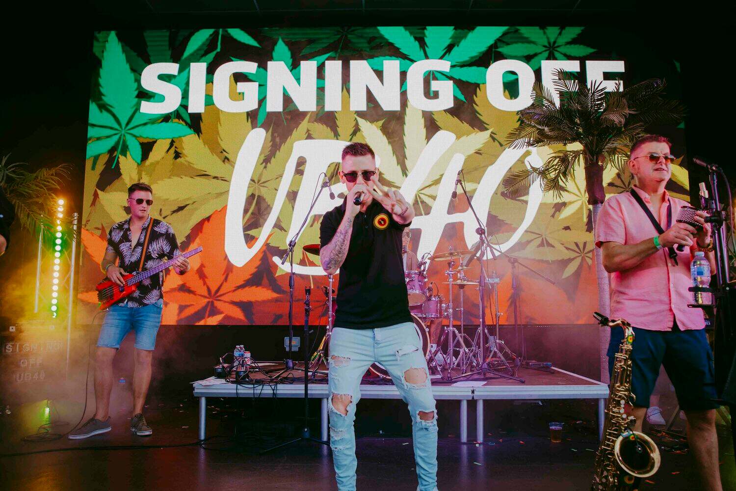 A band performs on stage with a large sign reading SIGNING OFF UB40 in the background. The lead singer holds a microphone, wearing sunglasses and ripped jeans. Other musicians play instruments, surrounded by colorful lights and a leafy backdrop.