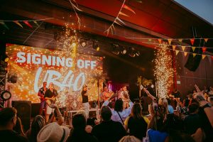A vibrant concert scene featuring a band performing on stage with Signing Off UB40 displayed on a large screen. Colorful lights, banners, and sparklers enhance the lively atmosphere. A crowd enthusiastically watches and enjoys the show.