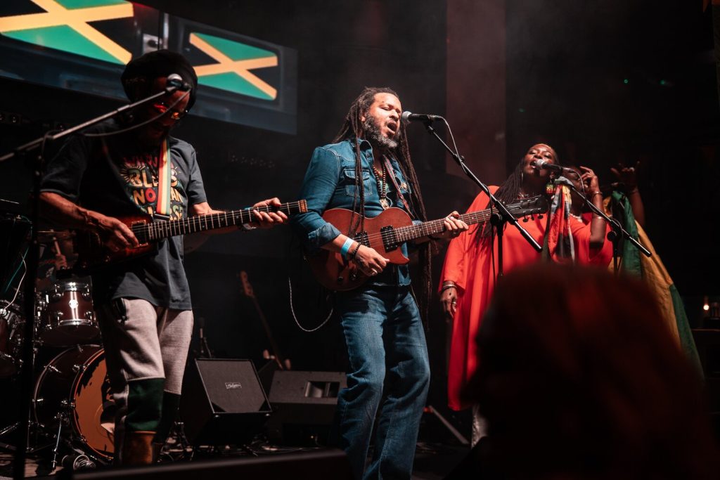 Three musicians perform on stage. The central figure plays an acoustic guitar and sings, while the others play and sing alongside. The background features a Jamaican flag. Stage lights create a vibrant atmosphere.