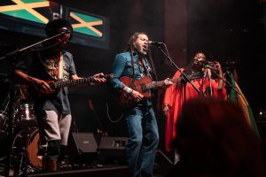 Three musicians perform on stage. The central figure plays an acoustic guitar and sings, while the others play and sing alongside. The background features a Jamaican flag. Stage lights create a vibrant atmosphere.