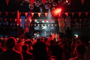 A band performs on stage in a lively nightclub. The venue is decorated with colorful flags and hanging disco balls. A crowd of people watches and dances under vibrant red and orange lights. Screens display digital visuals in the background.