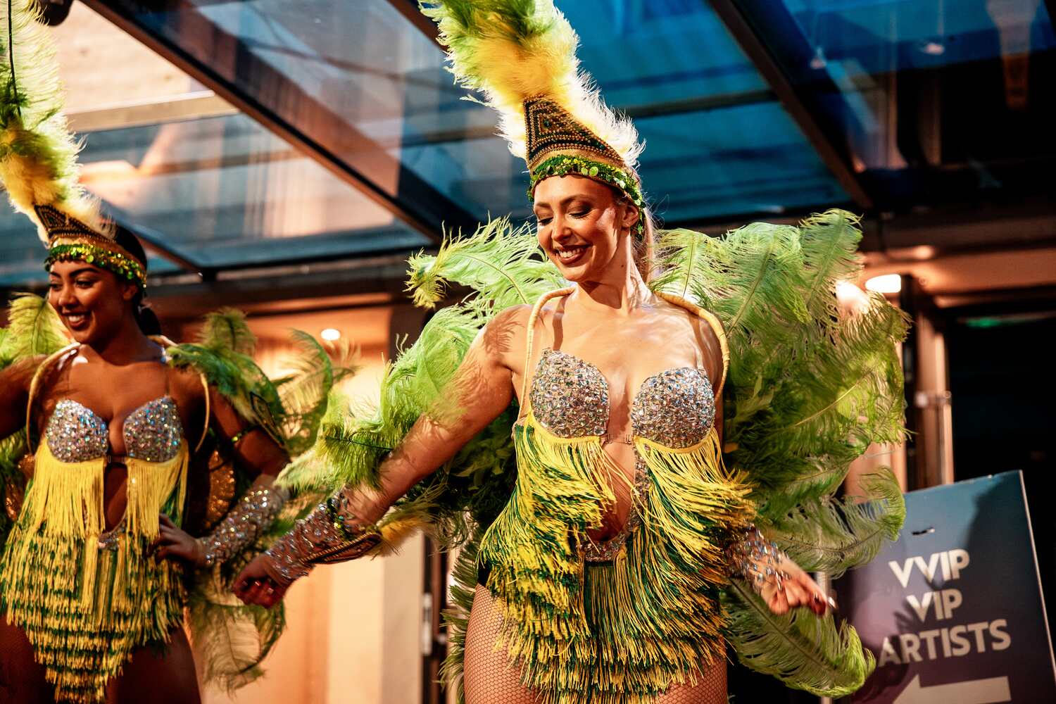 Dancers dressed in vibrant green and yellow feathered costumes with sparkling tops perform under a glass ceiling. They smile brightly as they move, with a sign in the background reading VVIP and VIP ARTISTS.