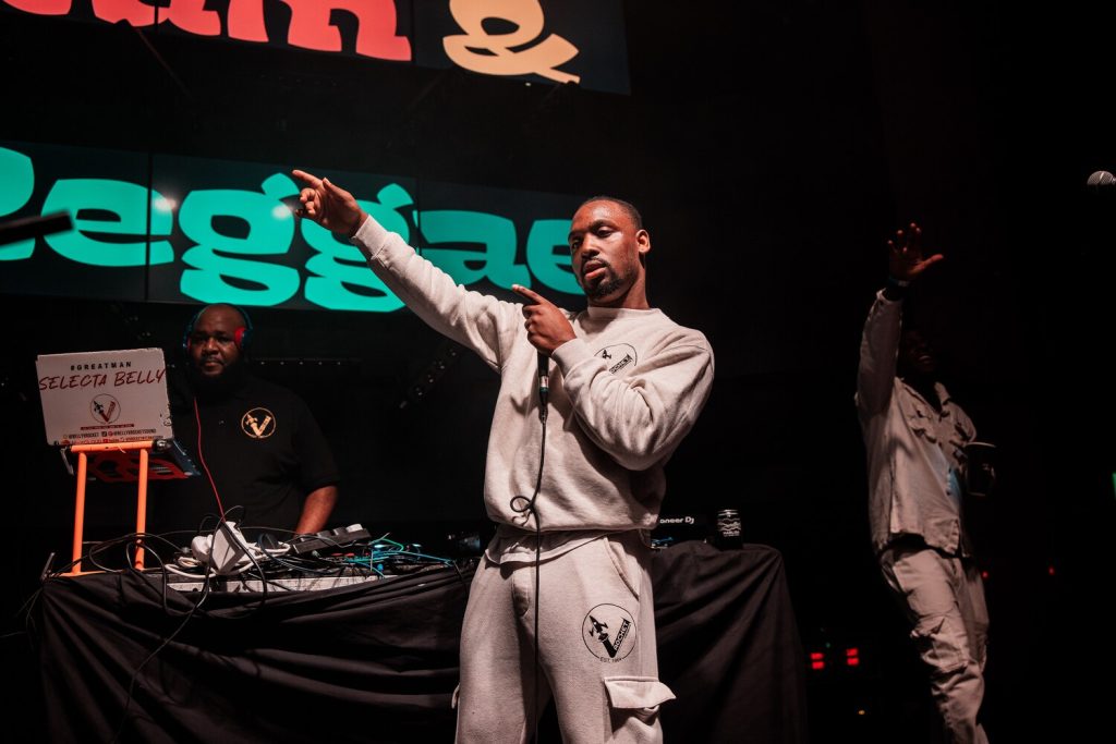 JackKimberPhotographyLtd_@jackkimberphoto-37 Man in a gray tracksuit holds a microphone and points while performing on stage. Another man stands near a DJ setup with a Selecta Belly sign. A third person is in the background with a hand raised. Bright lights and colorful signage are visible.