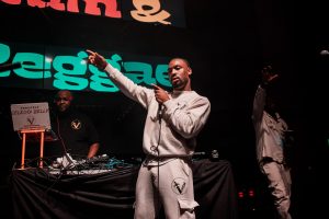Man in a gray tracksuit holds a microphone and points while performing on stage. Another man stands near a DJ setup with a Selecta Belly sign. A third person is in the background with a hand raised. Bright lights and colorful signage are visible.
