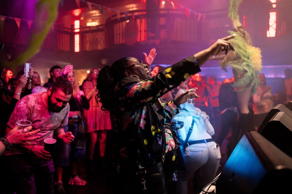 JackKimberPhotographyLtd_@jackkimberphoto-45 A lively concert scene with a crowd of people dancing and cheering. A person in a colorful jacket is energetically pointing towards the stage. Bright stage lighting and decorations create a vibrant atmosphere.