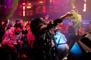 A lively concert scene with a crowd of people dancing and cheering. A person in a colorful jacket is energetically pointing towards the stage. Bright stage lighting and decorations create a vibrant atmosphere.